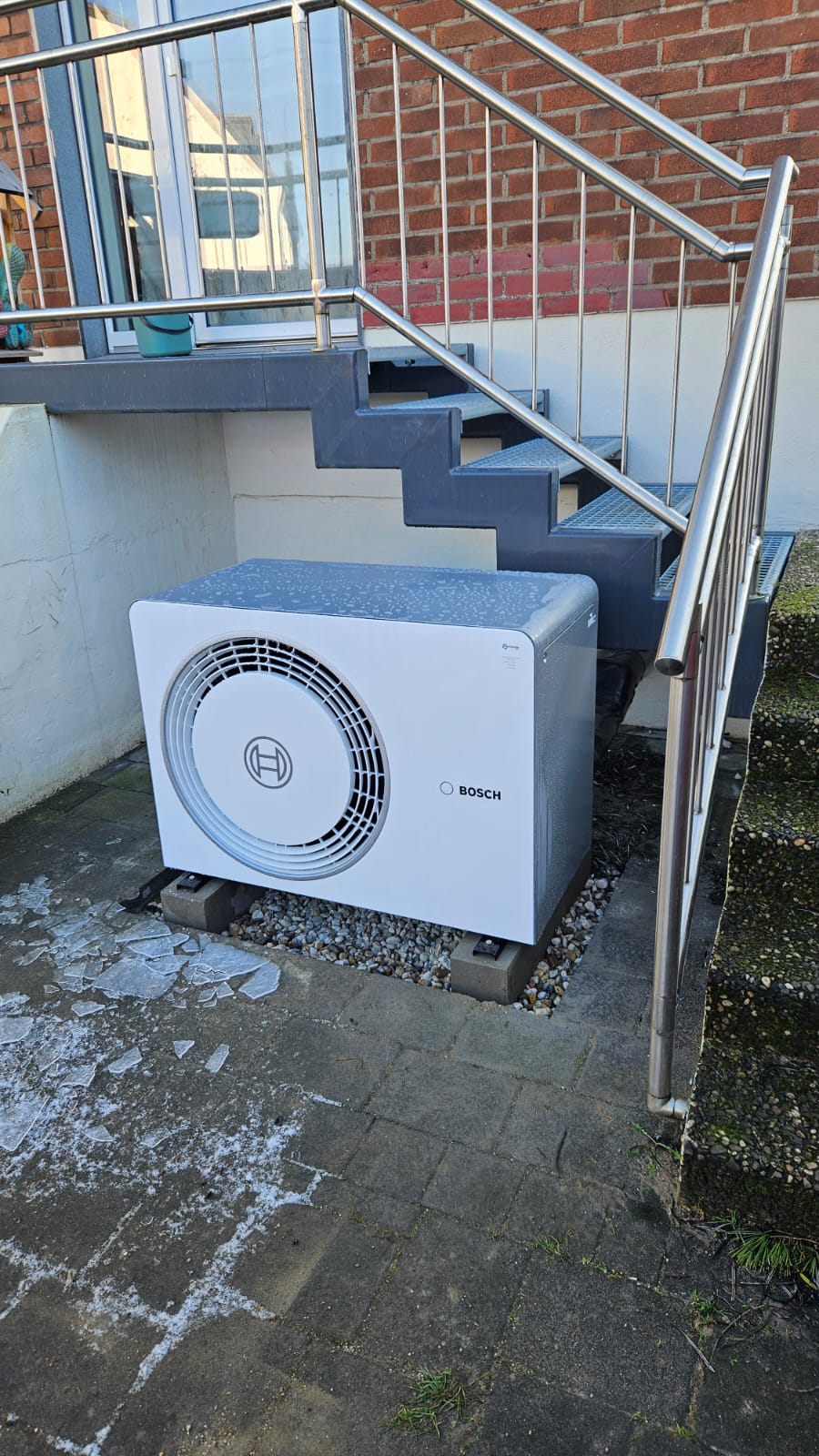 Bosch-Wärmepumpe im Außenbereich unter Treppe, umgeben von Eisplatten auf dem Boden – sichtbar nach Frost.
